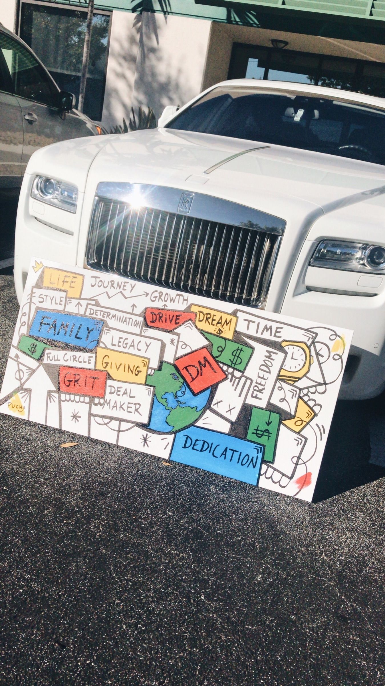 Colorful block painting with words like Family, Grit and Giving, leaning against a white Rolls-Royce