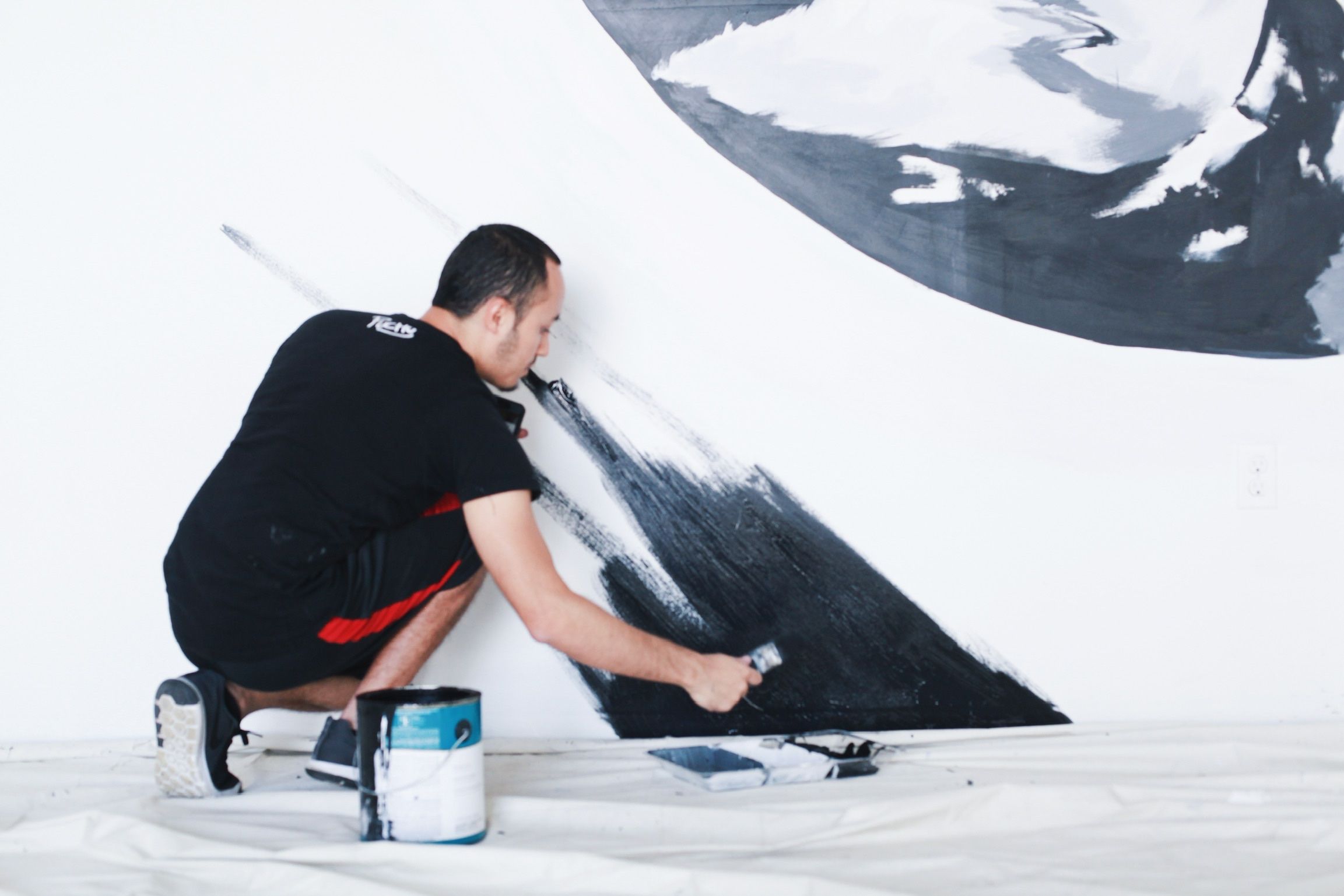 The artist crouching to paint a black shape on a white wall