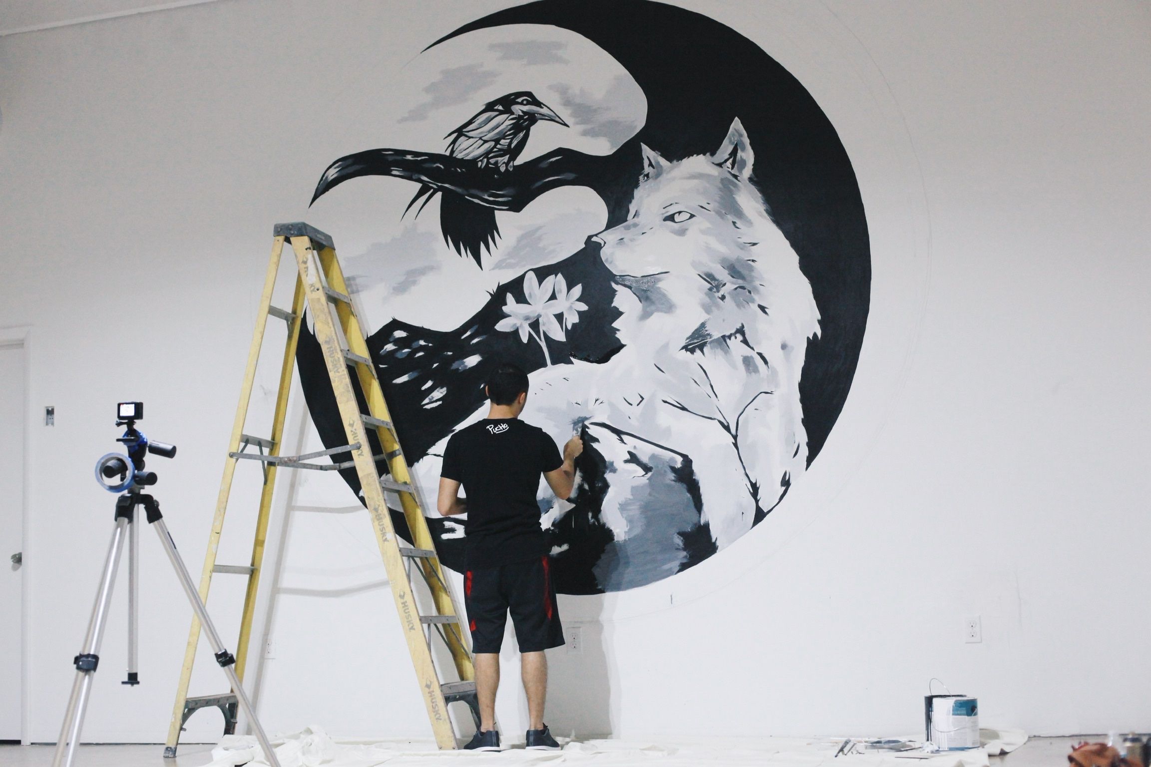 PUCHO painting a black and white mural of a wolf and a raven inside a crescent moon