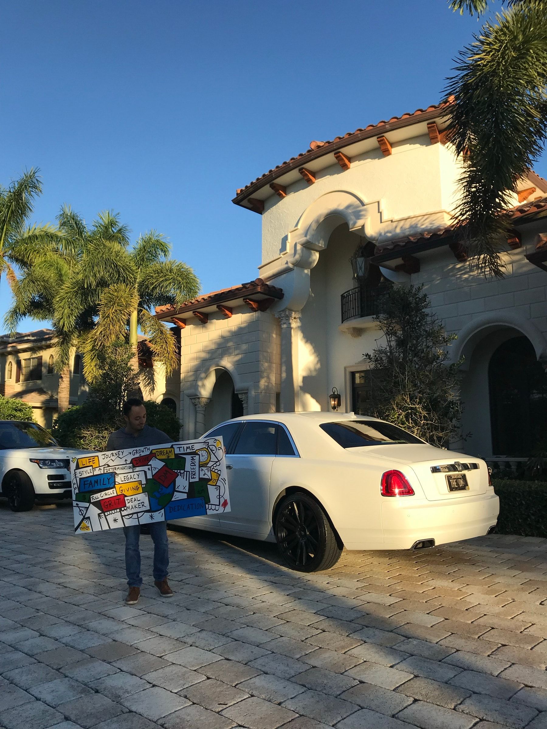 A collector holding a colorful word painting outside a Mediterranean home beside a white Rolls-Royce