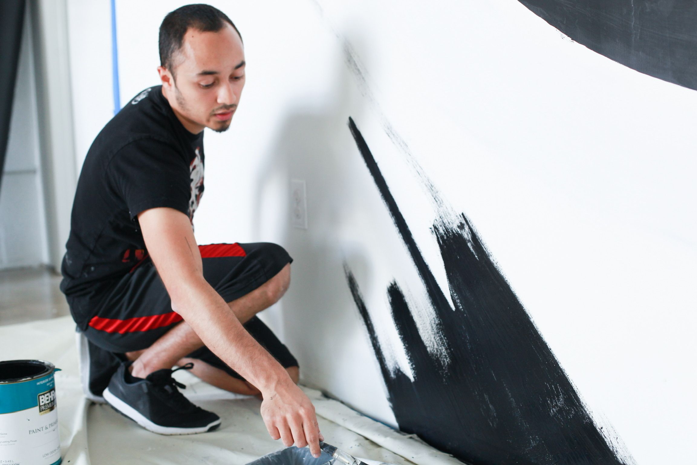 The artist kneeling beside paint cans, studying a black brushstroke mural